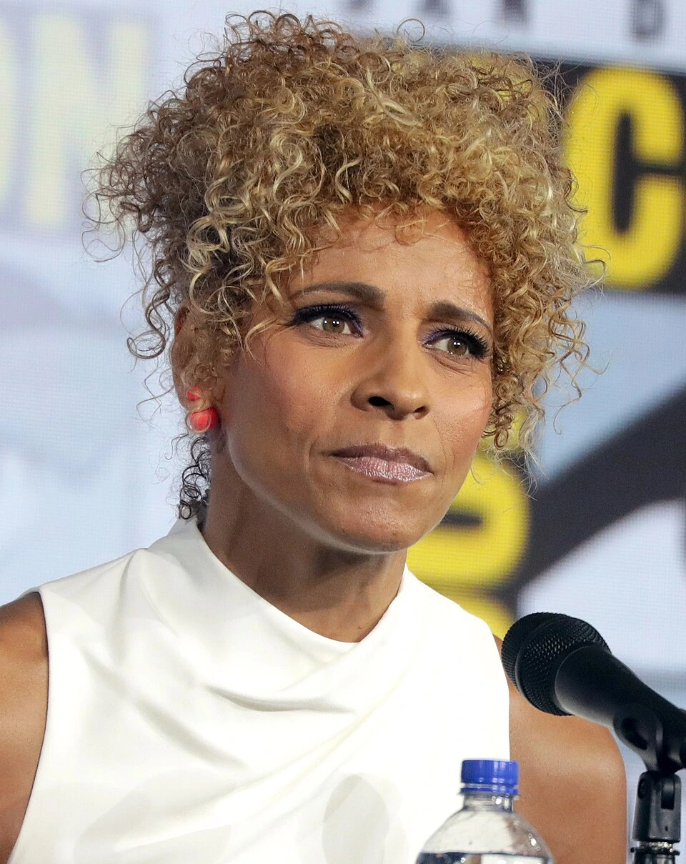 Michelle Hurd speaking at a public event, wearing a white top and standing at a microphone.