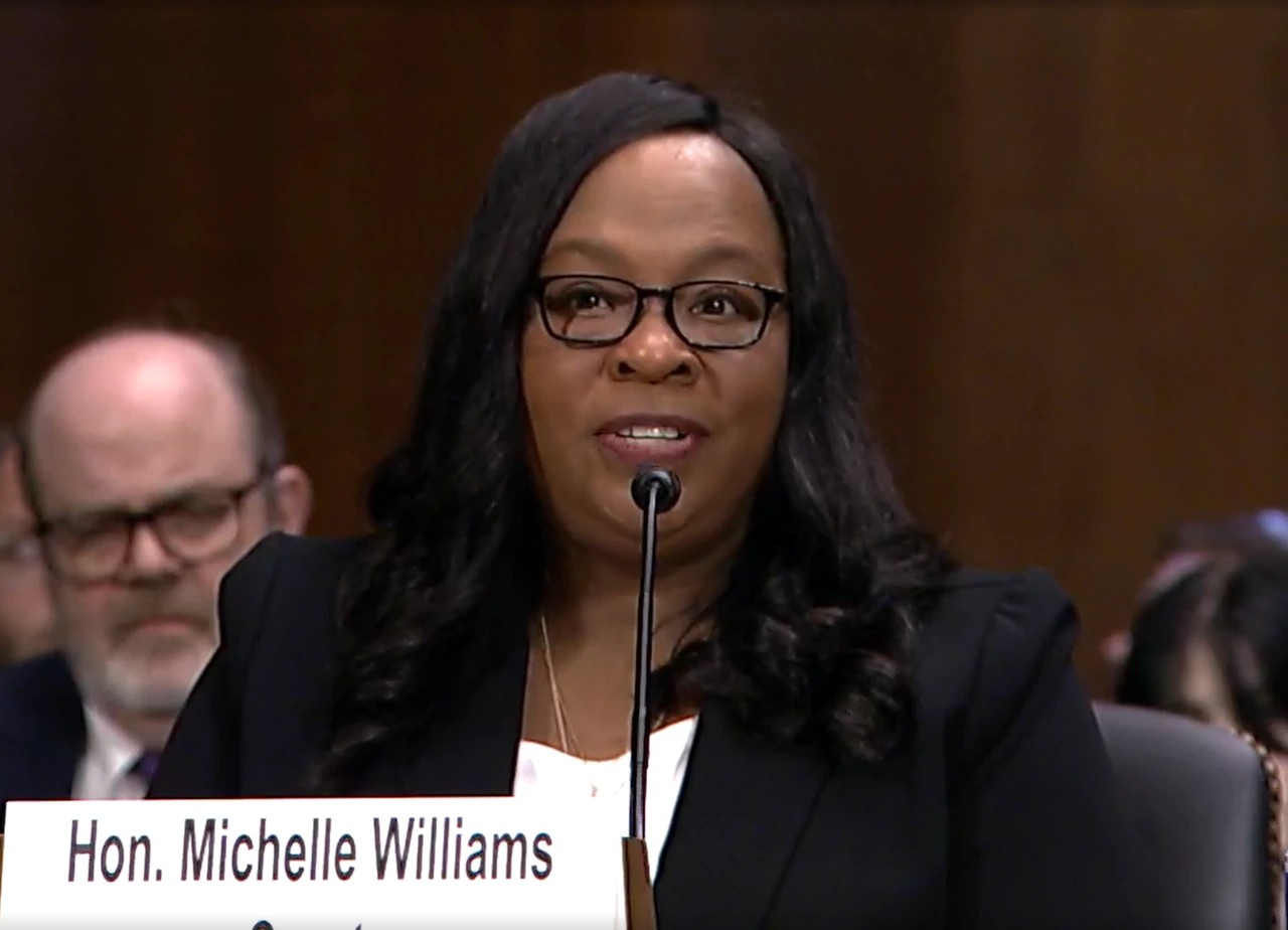 Michelle Williams speaking at a hearing with a name placard visible